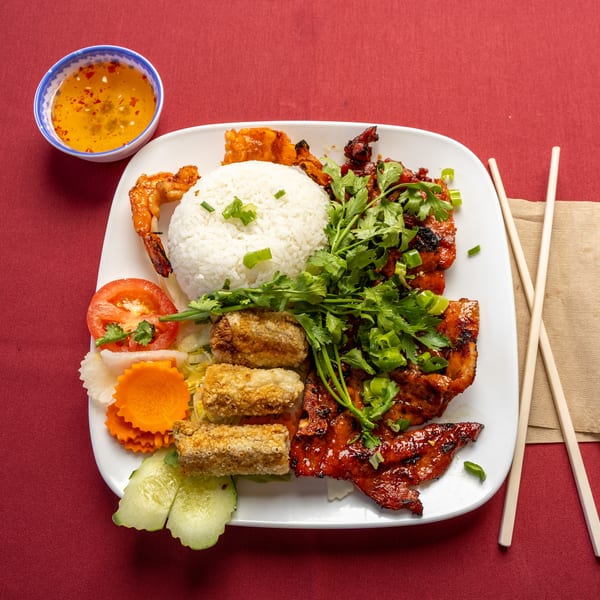Vietnamese grilled pork, shrimp, egg rolls, rice, and vegetables with dipping sauce on a red tablecloth.