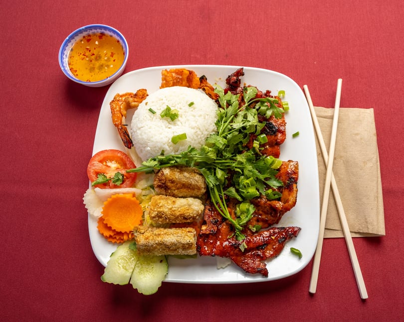 Vietnamese grilled pork, shrimp, egg rolls, rice, and vegetables with dipping sauce on a red tablecloth.