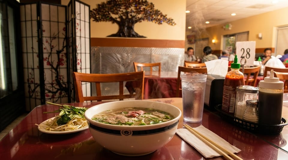 Steaming bowl of pho with bean sprouts and herbs on a table in a Vietnamese restaurant.