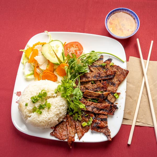 Grilled sliced beef with rice, salad, cilantro, and a dipping sauce on a red tablecloth.