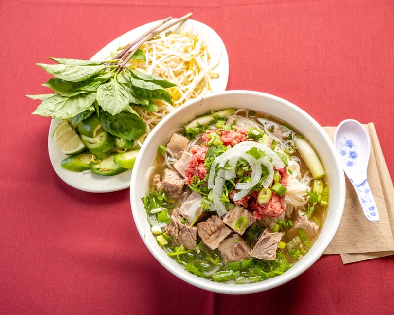 A bowl of pho with beef, noodles, and herbs, alongside a plate of bean sprouts, basil, and lime.